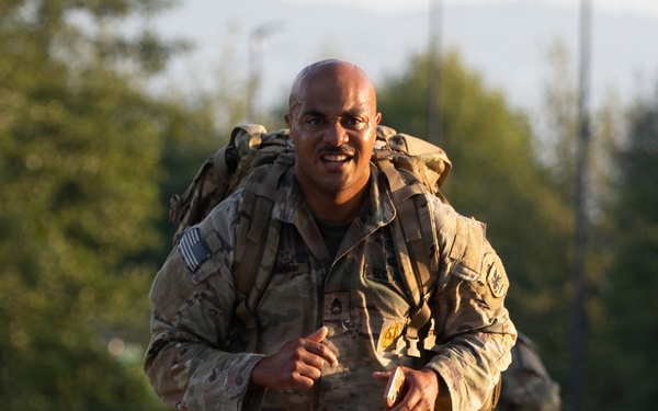 A U.S. Army soldier in Operational Camouflage Pattern (OCP) uniform is running or rucking (ruck march) along a paved road while carrying a large military rucksack. The soldier is smiling and appears to be in good spirits despite the physical exertion. A second soldier in similar uniform is visible in the background, also carrying a rucksack.