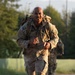A U.S. Army soldier in Operational Camouflage Pattern (OCP) uniform is running or rucking (ruck march) along a paved road while carrying a large military rucksack. The soldier is smiling and appears to be in good spirits despite the physical exertion. A second soldier in similar uniform is visible in the background, also carrying a rucksack.