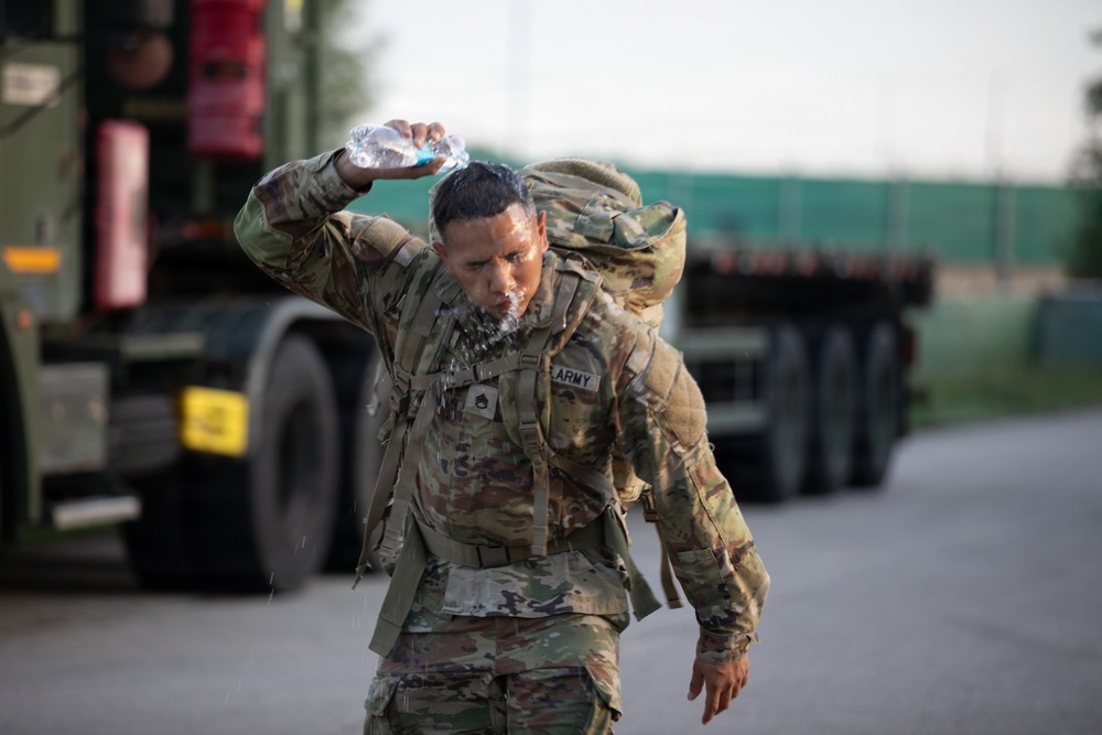A U.S. Army soldier in full combat uniform and rucksack is pouring water over their head, likely during a ruck march or physical training exercise. The soldier appears to be managing heat stress by cooling themselves with a water bottle while continuing to move. Military vehicles, including what appears to be a flatbed truck, are visible in the blurred background.