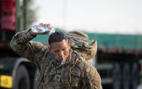 A U.S. Army soldier in full combat uniform and rucksack is pouring water over their head, likely during a ruck march or physical training exercise. The soldier appears to be managing heat stress by cooling themselves with a water bottle while continuing to move. Military vehicles, including what appears to be a flatbed truck, are visible in the blurred background.