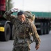 A U.S. Army soldier in full combat uniform and rucksack is pouring water over their head, likely during a ruck march or physical training exercise. The soldier appears to be managing heat stress by cooling themselves with a water bottle while continuing to move. Military vehicles, including what appears to be a flatbed truck, are visible in the blurred background.