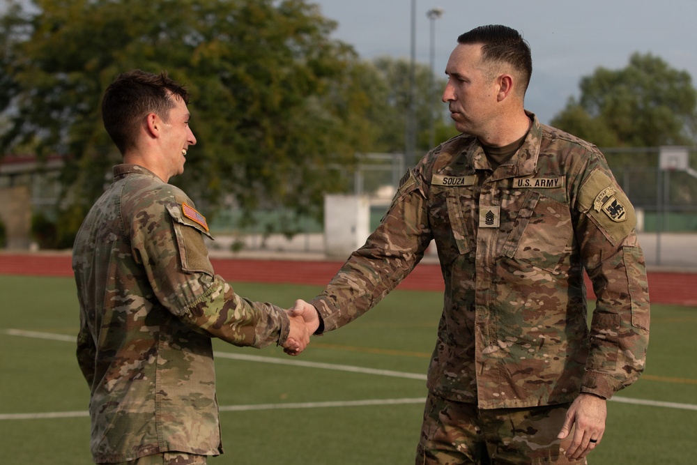 Two U.S. Army soldiers in OCP (Operational Camouflage Pattern) uniforms are shaking hands on what appears to be an athletic field or sports facility. The soldier on the right is identified by his name tape as 'SOUZA' and wears a Sergeant Major rank insignia, suggesting a formal recognition or greeting ceremony. The soldier on the left is smiling, indicating a positive interaction.