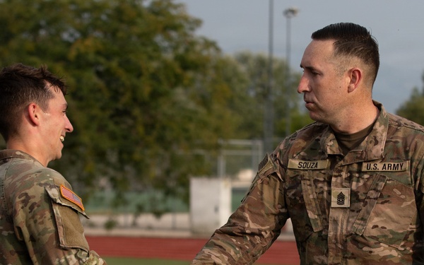 Two U.S. Army soldiers in OCP (Operational Camouflage Pattern) uniforms are shaking hands on what appears to be an athletic field or sports facility. The soldier on the right is identified by his name tape as 'SOUZA' and wears a Sergeant Major rank insignia, suggesting a formal recognition or greeting ceremony. The soldier on the left is smiling, indicating a positive interaction.