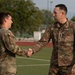 Two U.S. Army soldiers in OCP (Operational Camouflage Pattern) uniforms are shaking hands on what appears to be an athletic field or sports facility. The soldier on the right is identified by his name tape as 'SOUZA' and wears a Sergeant Major rank insignia, suggesting a formal recognition or greeting ceremony. The soldier on the left is smiling, indicating a positive interaction.