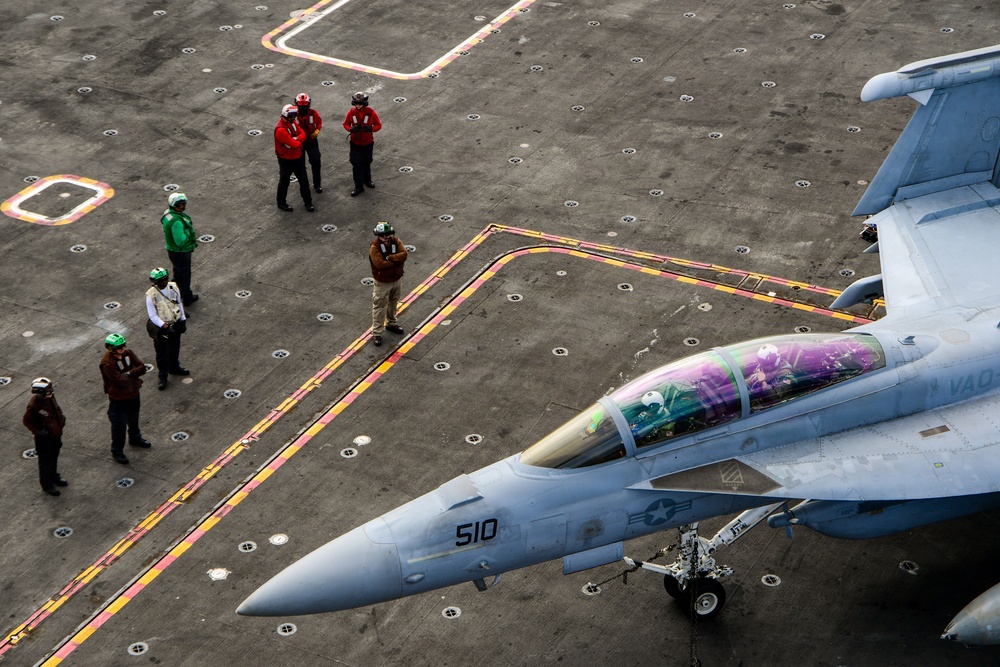 An aerial view of a U.S. Navy aircraft carrier flight deck showing a jet aircraft numbered '510' with 'VAQ' markings, indicating it is an electronic warfare aircraft (likely an EA-18G Growler). Multiple flight deck crew members in color-coded jerseys are visible standing on the deck near painted safety lines. The image is taken from an elevated angle, likely from the carrier's island superstructure.
