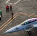 An aerial view of a U.S. Navy aircraft carrier flight deck showing a jet aircraft numbered '510' with 'VAQ' markings, indicating it is an electronic warfare aircraft (likely an EA-18G Growler). Multiple flight deck crew members in color-coded jerseys are visible standing on the deck near painted safety lines. The image is taken from an elevated angle, likely from the carrier's island superstructure.