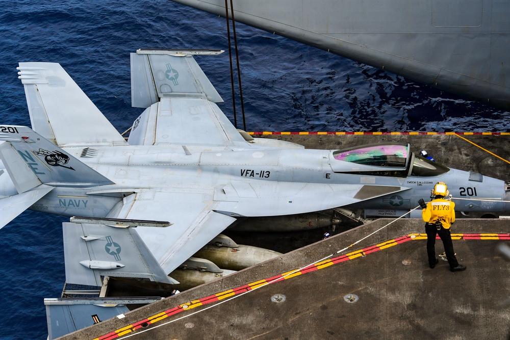 A U.S. Navy F/A-18E/F Super Hornet assigned to VFA-113 (the 'Stingers') is parked on the flight deck of an aircraft carrier, with its wings folded for deck storage. The aircraft bears the modex number 201 and tail code 'NF', indicating assignment to a carrier air wing. A flight deck crew member in a yellow jersey marked 'FLY 3' stands near the aircraft's nose, likely performing a supervisory or directing role.