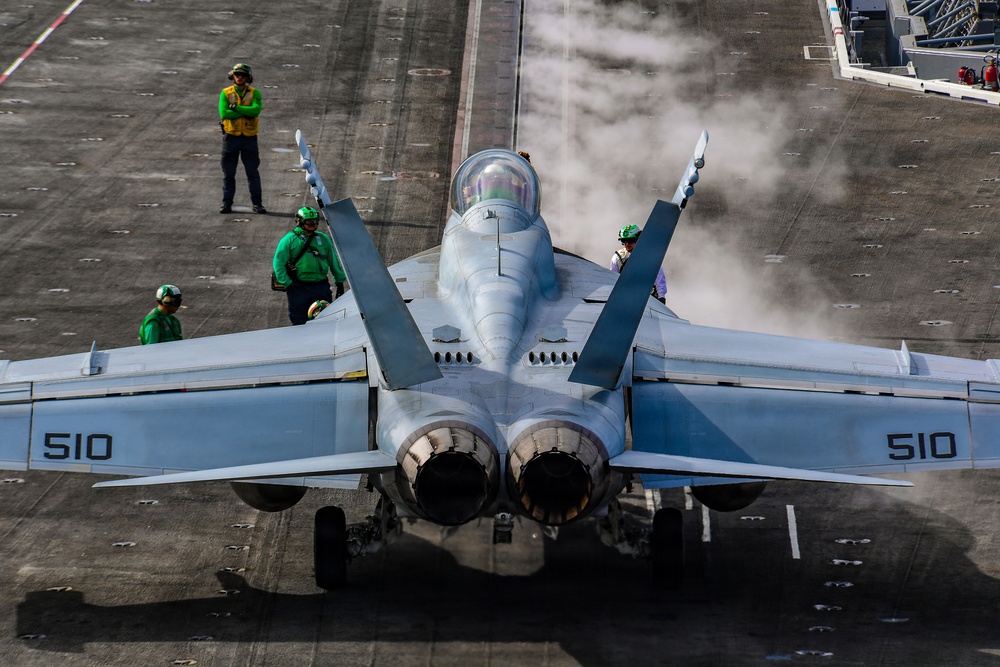 A U.S. Navy carrier-based jet aircraft, marked with the number 510 on both wings, is positioned on an aircraft carrier flight deck preparing for launch. Steam is visible rising from the catapult system in front of the aircraft. Several flight deck crew members in green jerseys and helmets are visible around the aircraft, performing pre-launch duties.