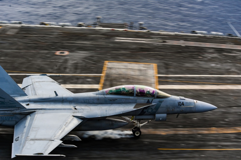 A U.S. Navy F/A-18 Super Hornet (bureau number 104) is captured during a catapult launch or high-speed takeoff roll on an aircraft carrier flight deck. The image shows significant motion blur in the background, indicating high speed, while the aircraft remains relatively sharp. The ocean is visible beyond the carrier deck, confirming the at-sea operational environment.