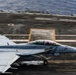 A U.S. Navy F/A-18 Super Hornet (bureau number 104) is captured during a catapult launch or high-speed takeoff roll on an aircraft carrier flight deck. The image shows significant motion blur in the background, indicating high speed, while the aircraft remains relatively sharp. The ocean is visible beyond the carrier deck, confirming the at-sea operational environment.