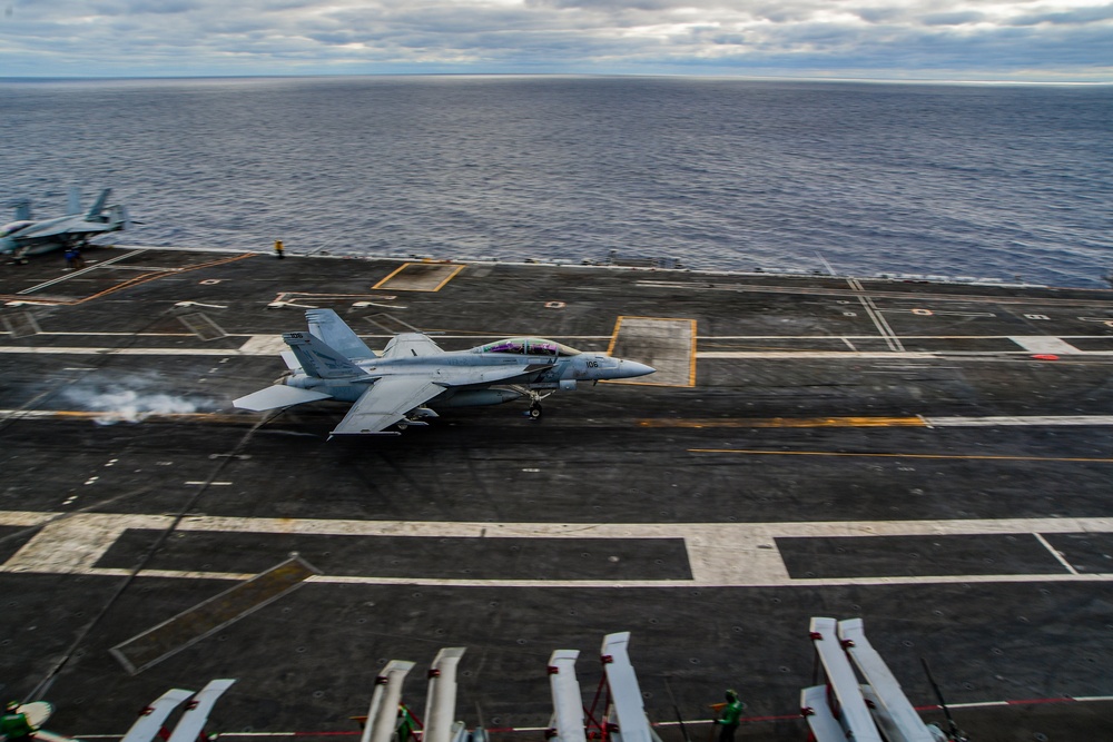 A U.S. Navy fighter jet, appearing to be an F/A-18 Super Hornet, is captured during a catapult launch or arrested landing on an aircraft carrier flight deck. The aircraft bears the modex number '406' and is producing exhaust smoke, indicating engine thrust. The carrier is underway on open ocean under overcast skies.