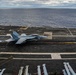 A U.S. Navy fighter jet, appearing to be an F/A-18 Super Hornet, is captured during a catapult launch or arrested landing on an aircraft carrier flight deck. The aircraft bears the modex number '406' and is producing exhaust smoke, indicating engine thrust. The carrier is underway on open ocean under overcast skies.