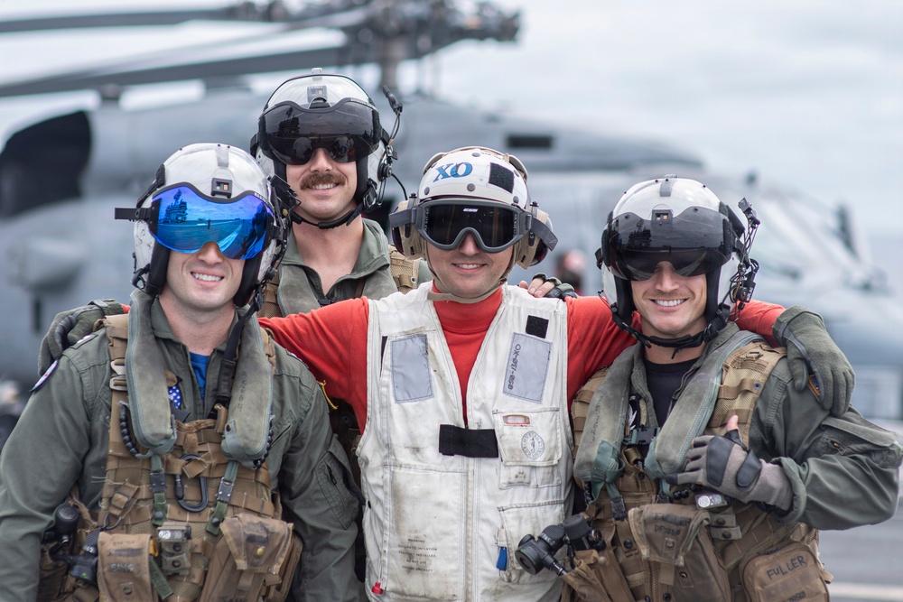 Four military personnel pose together on what appears to be a naval vessel flight deck, smiling for the camera. Three individuals are wearing flight suits with tactical gear and flight helmets, while the center individual wears a white deck vest and red shirt with 'XO' marked on their helmet. A military helicopter is visible in the blurred background.