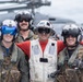 Four military personnel pose together on what appears to be a naval vessel flight deck, smiling for the camera. Three individuals are wearing flight suits with tactical gear and flight helmets, while the center individual wears a white deck vest and red shirt with 'XO' marked on their helmet. A military helicopter is visible in the blurred background.