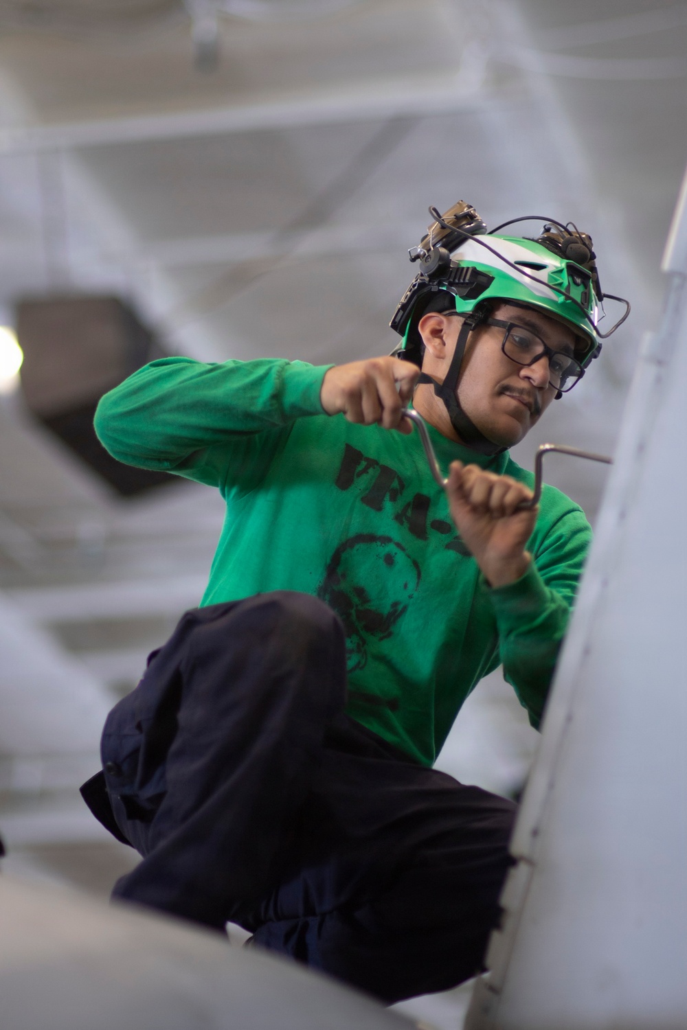 A young male technician wearing a green jersey/shirt with 'VFA-' text and a helmet equipped with night-vision goggle mounts is performing maintenance work. He is holding what appear to be safety wire or cotter pins in both hands, working on what looks like an aircraft surface or panel. The setting appears to be inside a hangar or aircraft maintenance bay.