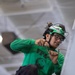 A young male technician wearing a green jersey/shirt with 'VFA-' text and a helmet equipped with night-vision goggle mounts is performing maintenance work. He is holding what appear to be safety wire or cotter pins in both hands, working on what looks like an aircraft surface or panel. The setting appears to be inside a hangar or aircraft maintenance bay.