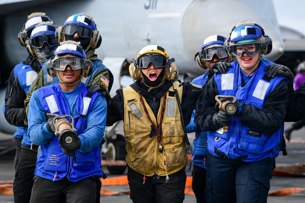 A group of U.S. Navy flight deck crew members are posed on an aircraft carrier flight deck, wearing color-coded safety gear indicating their roles. The central figure in a yellow vest appears to be shouting or cheering energetically, while others in blue vests stand alongside. A military aircraft with the number '511' is partially visible in the background.