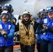 A group of U.S. Navy flight deck crew members are posed on an aircraft carrier flight deck, wearing color-coded safety gear indicating their roles. The central figure in a yellow vest appears to be shouting or cheering energetically, while others in blue vests stand alongside. A military aircraft with the number '511' is partially visible in the background.