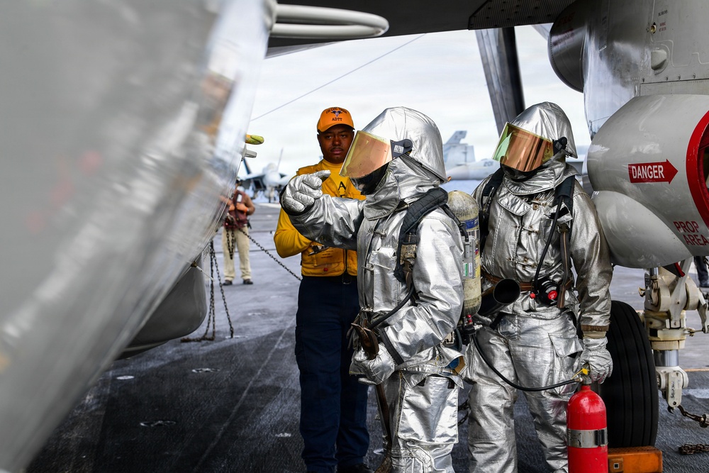The image shows personnel on what appears to be a naval aircraft carrier flight deck. Two individuals wearing silver aluminized proximity fire suits with reflective visors are being briefed or directed by a crew member in an orange vest and cap. A red fire extinguisher and aircraft components are visible in the foreground.