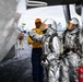The image shows personnel on what appears to be a naval aircraft carrier flight deck. Two individuals wearing silver aluminized proximity fire suits with reflective visors are being briefed or directed by a crew member in an orange vest and cap. A red fire extinguisher and aircraft components are visible in the foreground.