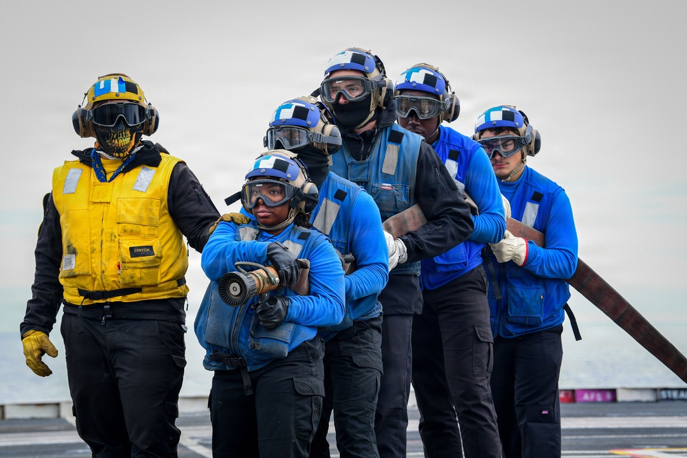 A group of military personnel, likely U.S. Navy sailors, are positioned on what appears to be the flight deck of an aircraft carrier. They are wearing color-coded gear consistent with naval flight deck operations, including blue jerseys/vests and one yellow vest, along with helmets, goggles, and gloves. The team appears to be conducting a firefighting drill or damage control exercise, with the front personnel holding a fire hose nozzle.