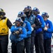 A group of military personnel, likely U.S. Navy sailors, are positioned on what appears to be the flight deck of an aircraft carrier. They are wearing color-coded gear consistent with naval flight deck operations, including blue jerseys/vests and one yellow vest, along with helmets, goggles, and gloves. The team appears to be conducting a firefighting drill or damage control exercise, with the front personnel holding a fire hose nozzle.