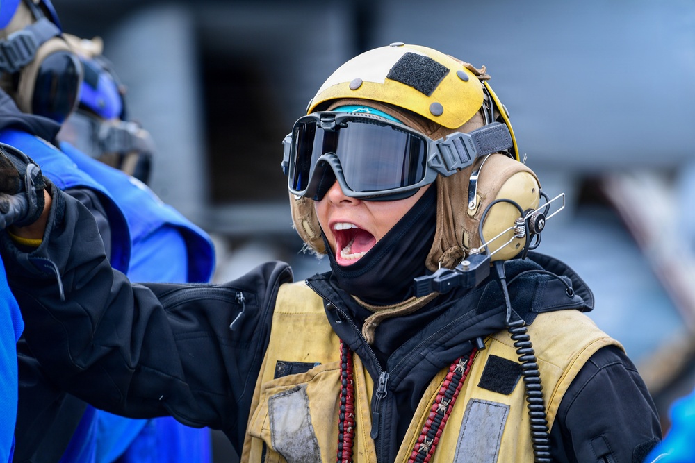 A person wearing military or naval flight deck crew equipment, including a yellow helmet, tinted goggles, a communications headset, and a tan/yellow tactical vest, appears to be shouting or giving a vocal command. The individual is dressed in cold-weather gear with a black balaclava and jacket underneath. The setting appears to be an outdoor operational environment, possibly a ship flight deck or military installation.