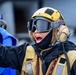 A person wearing military or naval flight deck crew equipment, including a yellow helmet, tinted goggles, a communications headset, and a tan/yellow tactical vest, appears to be shouting or giving a vocal command. The individual is dressed in cold-weather gear with a black balaclava and jacket underneath. The setting appears to be an outdoor operational environment, possibly a ship flight deck or military installation.