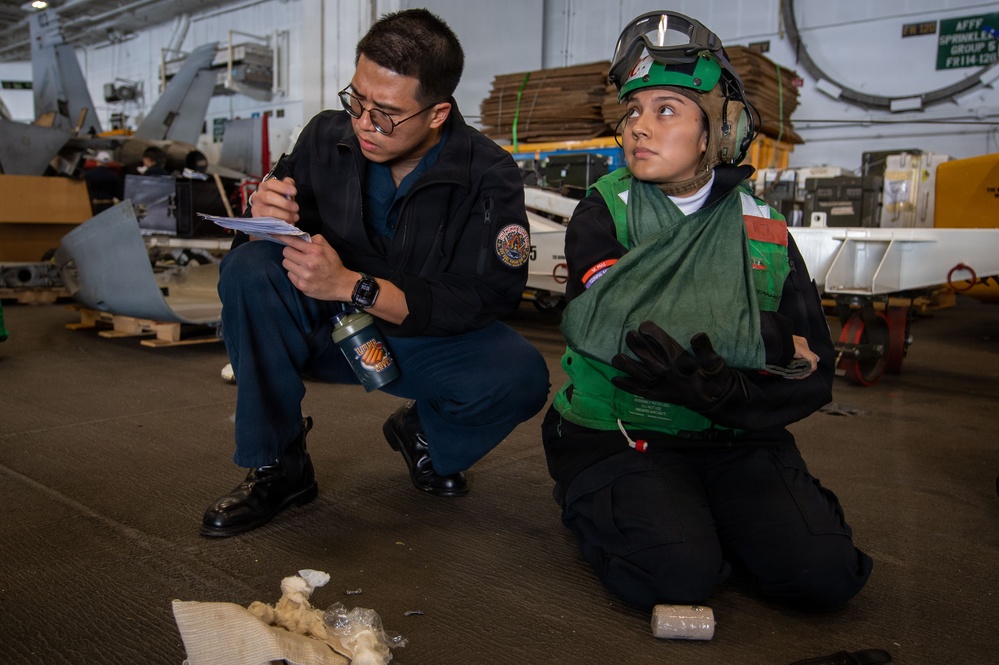 Two military personnel, likely U.S. Navy sailors, are conducting what appears to be a training exercise or inspection in a hangar bay aboard a naval vessel. One sailor in navy blue coveralls is crouching and writing on a clipboard, while the other is kneeling and wearing a green flight deck vest, helmet with goggles, and hearing protection. Various aircraft components and equipment are visible in the background.