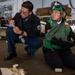 Two military personnel, likely U.S. Navy sailors, are conducting what appears to be a training exercise or inspection in a hangar bay aboard a naval vessel. One sailor in navy blue coveralls is crouching and writing on a clipboard, while the other is kneeling and wearing a green flight deck vest, helmet with goggles, and hearing protection. Various aircraft components and equipment are visible in the background.