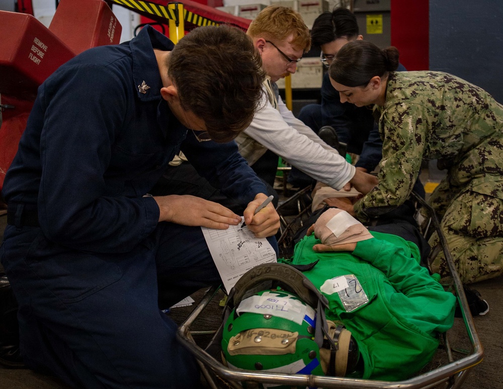 Military personnel, including Navy sailors in NWU (Navy Working Uniform) camouflage and dark blue coveralls, are conducting a medical training exercise or drill. A simulated casualty in a green flight suit is lying on a stretcher/litter, with personnel documenting information on a form and attending to the mannequin or simulated patient. The setting appears to be inside a naval vessel, likely an aircraft carrier hangar bay, indicated by the 'REMOVE BEFORE FLIGHT' tag visible in the background.