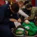 Military personnel, including Navy sailors in NWU (Navy Working Uniform) camouflage and dark blue coveralls, are conducting a medical training exercise or drill. A simulated casualty in a green flight suit is lying on a stretcher/litter, with personnel documenting information on a form and attending to the mannequin or simulated patient. The setting appears to be inside a naval vessel, likely an aircraft carrier hangar bay, indicated by the 'REMOVE BEFORE FLIGHT' tag visible in the background.