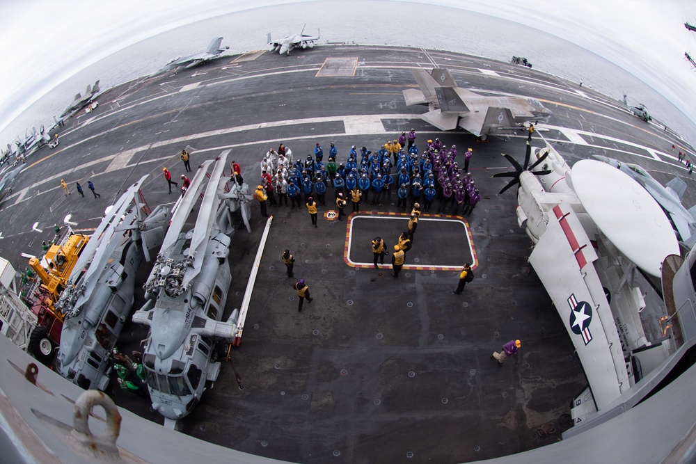 A fisheye aerial photograph taken from an elevated position on a US Navy aircraft carrier flight deck, showing a large group of personnel assembled in color-coded jerseys around a helicopter landing spot. Multiple aircraft are visible on deck including helicopters and what appears to be a fixed-wing aircraft, suggesting a formal muster or ceremony is taking place. The wide-angle lens distortion gives a curved horizon with open ocean visible in the background.