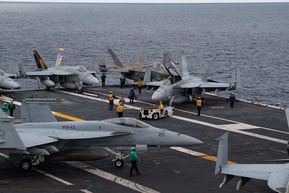 The image shows the flight deck of a U.S. Navy aircraft carrier at sea, with multiple fixed-wing aircraft staged and being repositioned by deck crew. At least one F/A-18 Super Hornet marked 'VFA-113' with modex '211' is prominently visible in the foreground, along with what appears to be an F-35C in the middle distance. Deck personnel in color-coded jerseys are actively conducting flight deck operations.