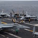 The image shows the flight deck of a U.S. Navy aircraft carrier at sea, with multiple fixed-wing aircraft staged and being repositioned by deck crew. At least one F/A-18 Super Hornet marked 'VFA-113' with modex '211' is prominently visible in the foreground, along with what appears to be an F-35C in the middle distance. Deck personnel in color-coded jerseys are actively conducting flight deck operations.