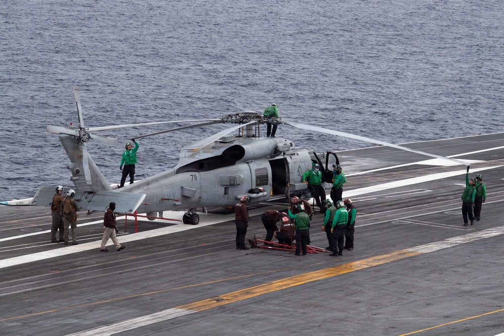 A military helicopter, marked with the number '711', is on the flight deck of an aircraft carrier surrounded by deck crew in green jerseys performing maintenance or pre-flight operations. The ocean is visible in the background, confirming the carrier is at sea. Multiple personnel are engaged in various tasks around and on top of the helicopter.