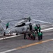 A military helicopter, marked with the number '711', is on the flight deck of an aircraft carrier surrounded by deck crew in green jerseys performing maintenance or pre-flight operations. The ocean is visible in the background, confirming the carrier is at sea. Multiple personnel are engaged in various tasks around and on top of the helicopter.