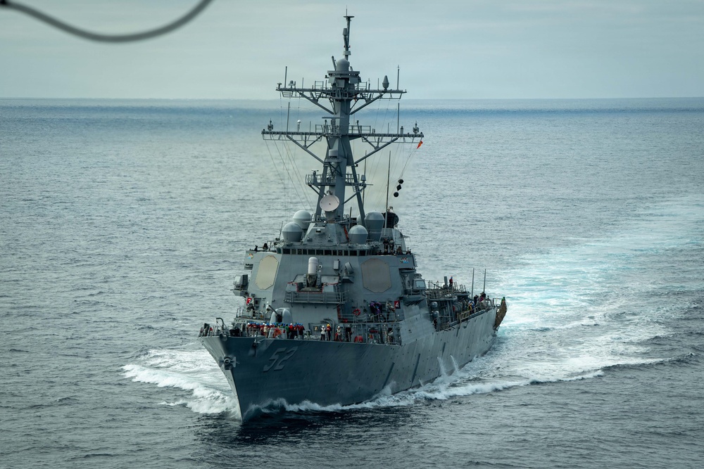 A U.S. Navy Arleigh Burke-class guided-missile destroyer is underway at sea, viewed from a slightly elevated forward angle. The hull number '52' is partially visible on the bow, and crew members can be seen on deck. The vessel is cutting through calm open ocean waters under an overcast sky.