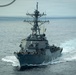 A U.S. Navy Arleigh Burke-class guided-missile destroyer is underway at sea, viewed from a slightly elevated forward angle. The hull number '52' is partially visible on the bow, and crew members can be seen on deck. The vessel is cutting through calm open ocean waters under an overcast sky.