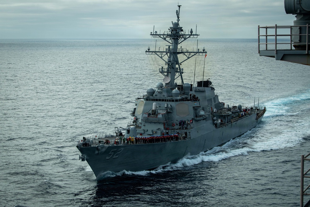 A U.S. Navy Arleigh Burke-class guided-missile destroyer bearing hull number 52 is underway at sea, photographed from an elevated position on a nearby vessel. The ship's crew is manning the rails in dress formation along the port side, suggesting a formal naval ceremony or port approach. The vessel is cutting through calm, overcast ocean waters leaving a visible wake.