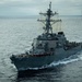 A U.S. Navy Arleigh Burke-class guided-missile destroyer bearing hull number 52 is underway at sea, photographed from an elevated position on a nearby vessel. The ship's crew is manning the rails in dress formation along the port side, suggesting a formal naval ceremony or port approach. The vessel is cutting through calm, overcast ocean waters leaving a visible wake.