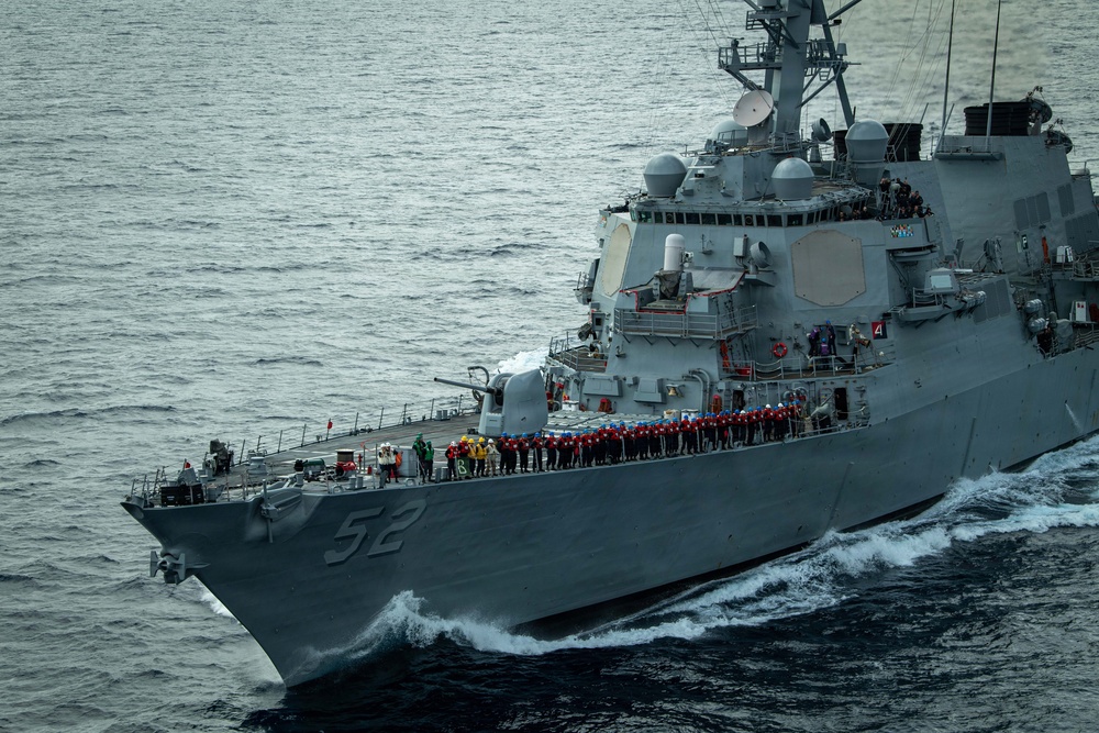 A U.S. Navy Arleigh Burke-class guided-missile destroyer bearing hull number 52 is underway at sea. A large formation of crew members in red hard hats are lined up along the main deck in what appears to be a manning-the-rails ceremony or muster. The vessel is cutting through choppy gray waters at moderate speed, with bow waves visible.