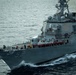 A U.S. Navy Arleigh Burke-class guided-missile destroyer bearing hull number 52 is underway at sea. A large formation of crew members in red hard hats are lined up along the main deck in what appears to be a manning-the-rails ceremony or muster. The vessel is cutting through choppy gray waters at moderate speed, with bow waves visible.