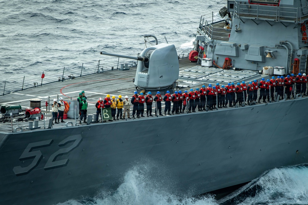 A U.S. Navy destroyer bearing hull number 52 is underway at sea with crew members manning the rails in a formal formation. Personnel are wearing red life vests and blue hard hats, consistent with an underway replenishment (UNREP) or special sea detail evolution. The ship features a forward 5-inch deck gun mount and vertical launch system (VLS) cells visible on the foredeck.