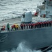 A U.S. Navy destroyer bearing hull number 52 is underway at sea with crew members manning the rails in a formal formation. Personnel are wearing red life vests and blue hard hats, consistent with an underway replenishment (UNREP) or special sea detail evolution. The ship features a forward 5-inch deck gun mount and vertical launch system (VLS) cells visible on the foredeck.