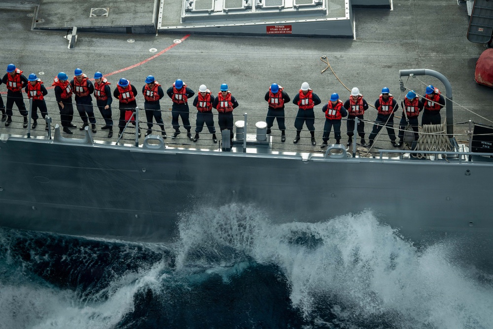 An aerial/elevated view of a naval vessel underway at sea, showing a line of approximately 14 sailors standing at the ship's rail. The personnel are wearing red life vests and blue or white hard hats, suggesting a formal muster, man-overboard drill, or replenishment-at-sea (RAS) evolution. The ship is cutting through choppy ocean water, producing visible white wake and spray along the hull.