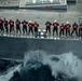 An aerial/elevated view of a naval vessel underway at sea, showing a line of approximately 14 sailors standing at the ship's rail. The personnel are wearing red life vests and blue or white hard hats, suggesting a formal muster, man-overboard drill, or replenishment-at-sea (RAS) evolution. The ship is cutting through choppy ocean water, producing visible white wake and spray along the hull.