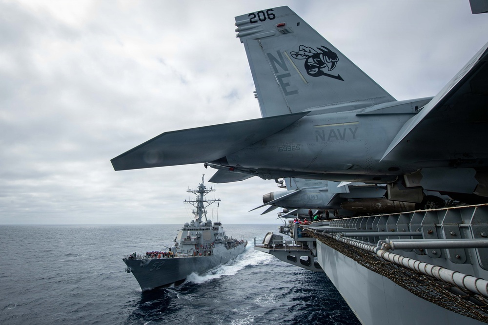 A U.S. Navy F/A-18 Super Hornet aircraft (modex 206, serial 169965) is parked on the flight deck edge of an aircraft carrier, with tail code 'NE' indicating assignment to a Pacific Fleet air wing. A U.S. Navy Arleigh Burke-class guided-missile destroyer is sailing in close formation alongside the carrier. The scene appears to be an underway replenishment or formation sailing operation in overcast open ocean conditions.