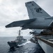 A U.S. Navy F/A-18 Super Hornet aircraft (modex 206, serial 169965) is parked on the flight deck edge of an aircraft carrier, with tail code 'NE' indicating assignment to a Pacific Fleet air wing. A U.S. Navy Arleigh Burke-class guided-missile destroyer is sailing in close formation alongside the carrier. The scene appears to be an underway replenishment or formation sailing operation in overcast open ocean conditions.