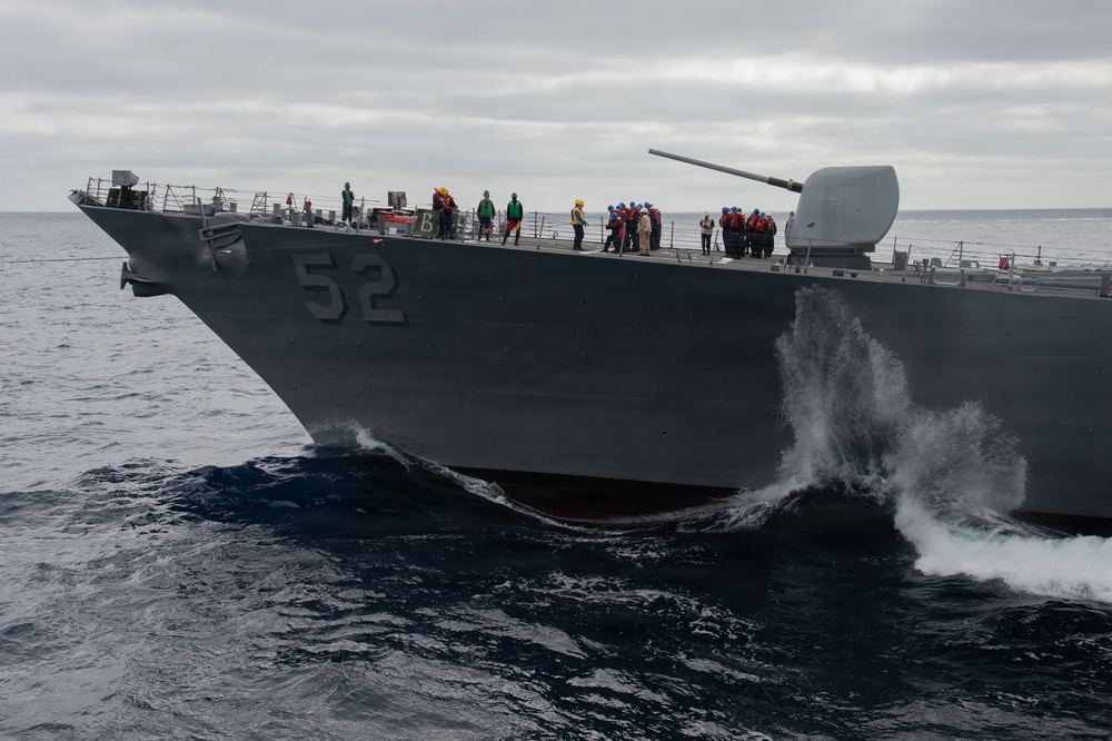 A U.S. Navy warship bearing hull number 52 is underway at sea, cutting through dark blue ocean waters with visible bow waves and spray. Multiple crew members in colored safety gear are visible on the deck, and a naval gun turret is mounted on the bow. The overcast sky and choppy seas suggest open ocean conditions.